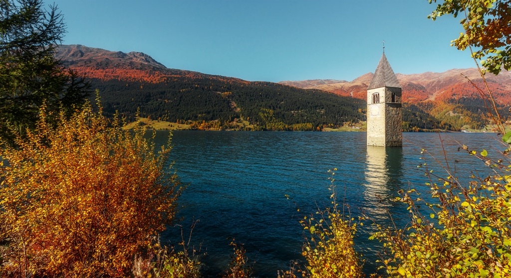Der versunkene Kirchturm im Reschensee vor den herbstlichen Hängen des Oberen Vinschgaus