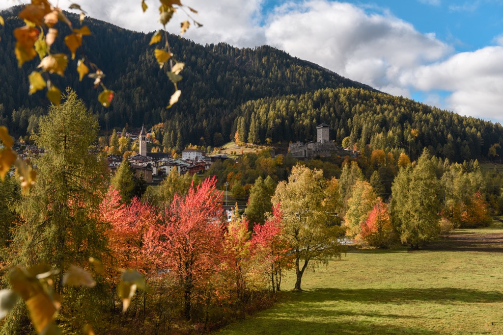 Ossana im Val di Sole im Herbst, mit Burg und Kirche unter einem Fichtenhang voller gelber Lärchen