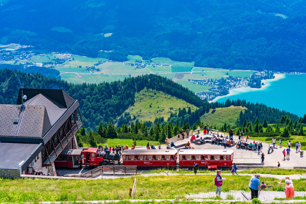 Red carriages of the Schafberg rack railway at the summit station, with crowds of visitors on the terraces and Wolfgangsee far below