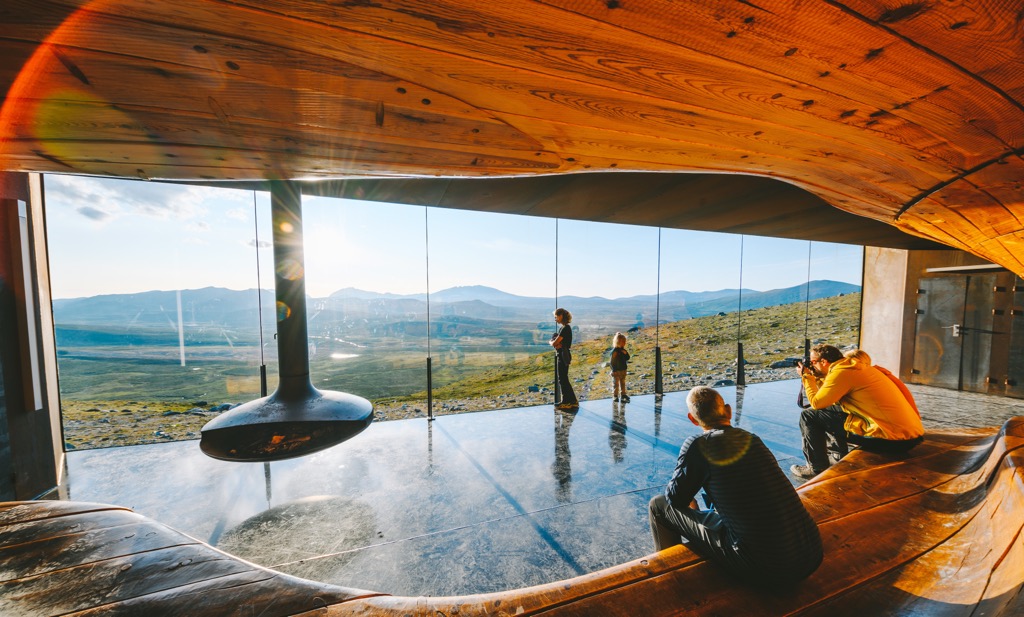 Inside Tverrfjellhytta: visitors on the curved wooden bench photograph the plateau through the glass wall, beside a hanging fireplace