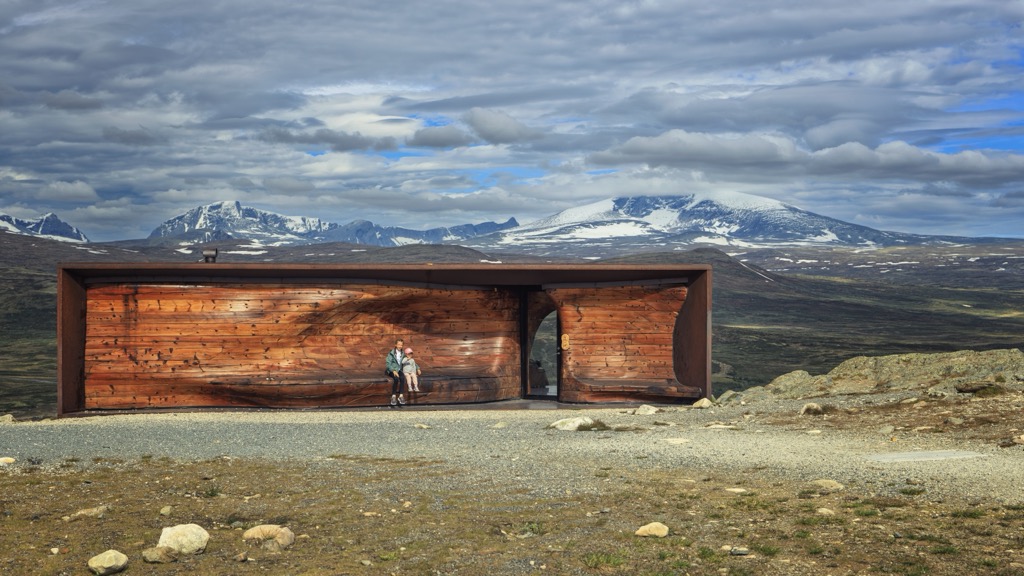 The long low Tverrfjellhytta pavilion on the open Dovrefjell plateau, with two visitors on its curved wooden bench and the snowy Snøhetta massif behind